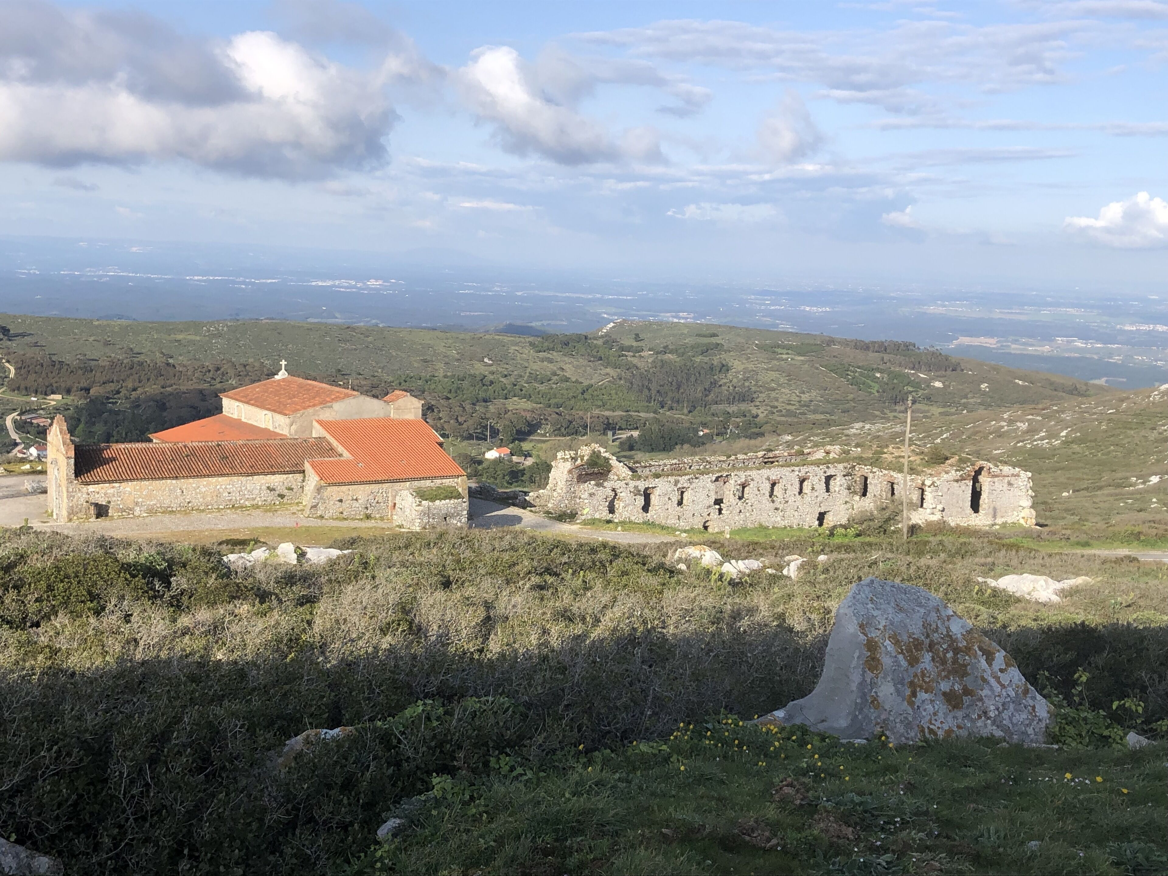Un bâtiment historique, doté d’un toit recouvert de tuiles rouges et de murs en pierre, entouré de verdure et offrant une vue sur un paysage vallonné.