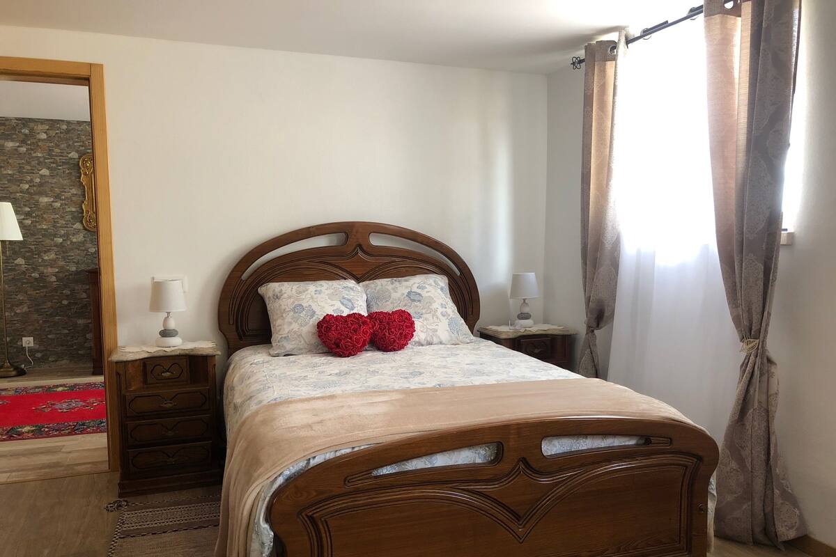 A neatly made bed with a wooden headboard, two bedside lamps, and a decorative heart pillow.