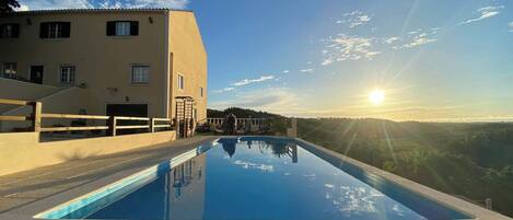 A swimming pool with a clear view of the sunset, a building with a sloped roof, and a paved deck.