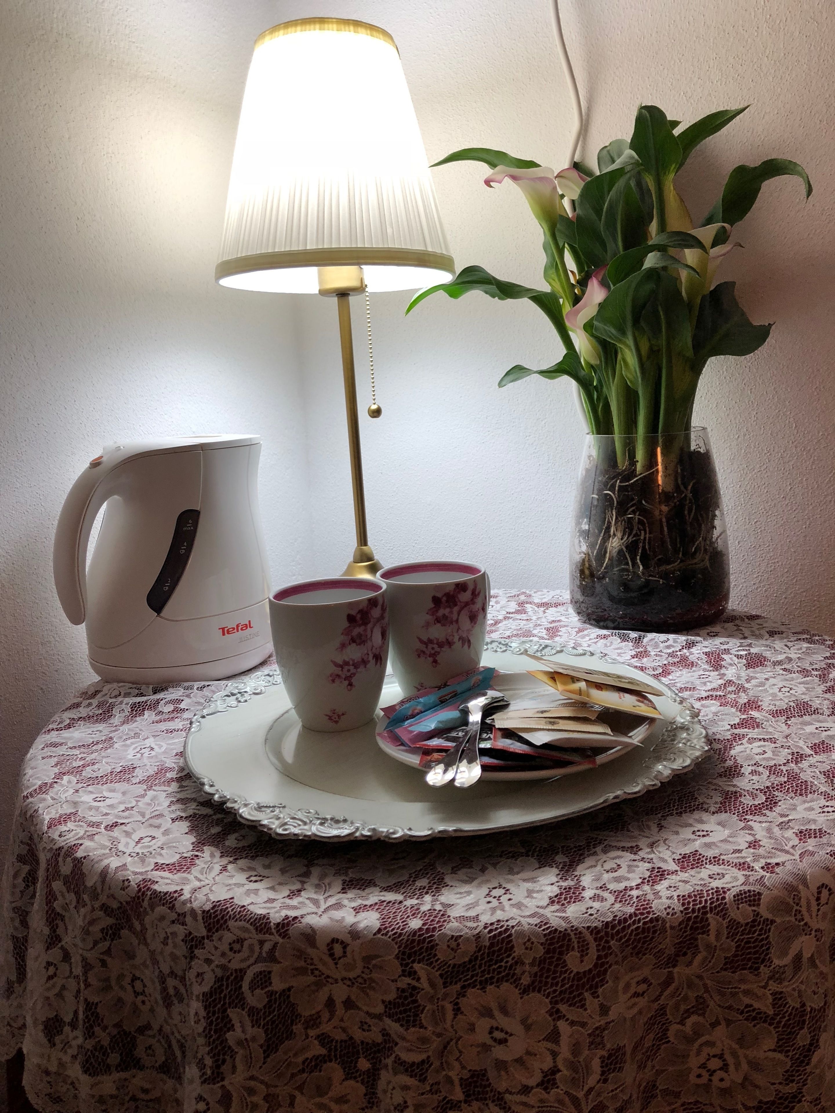 A table with a lace tablecloth, a white ceramic pitcher, two floral cups, a tray with a spoon and packets, a vase with green plants, and a table lamp.