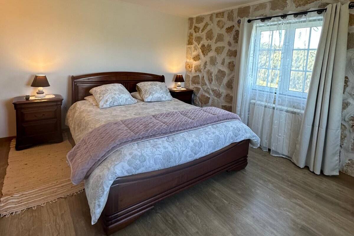 A bedroom with a wooden bed, two bedside tables with lamps, a stone wall, and a window with curtains.