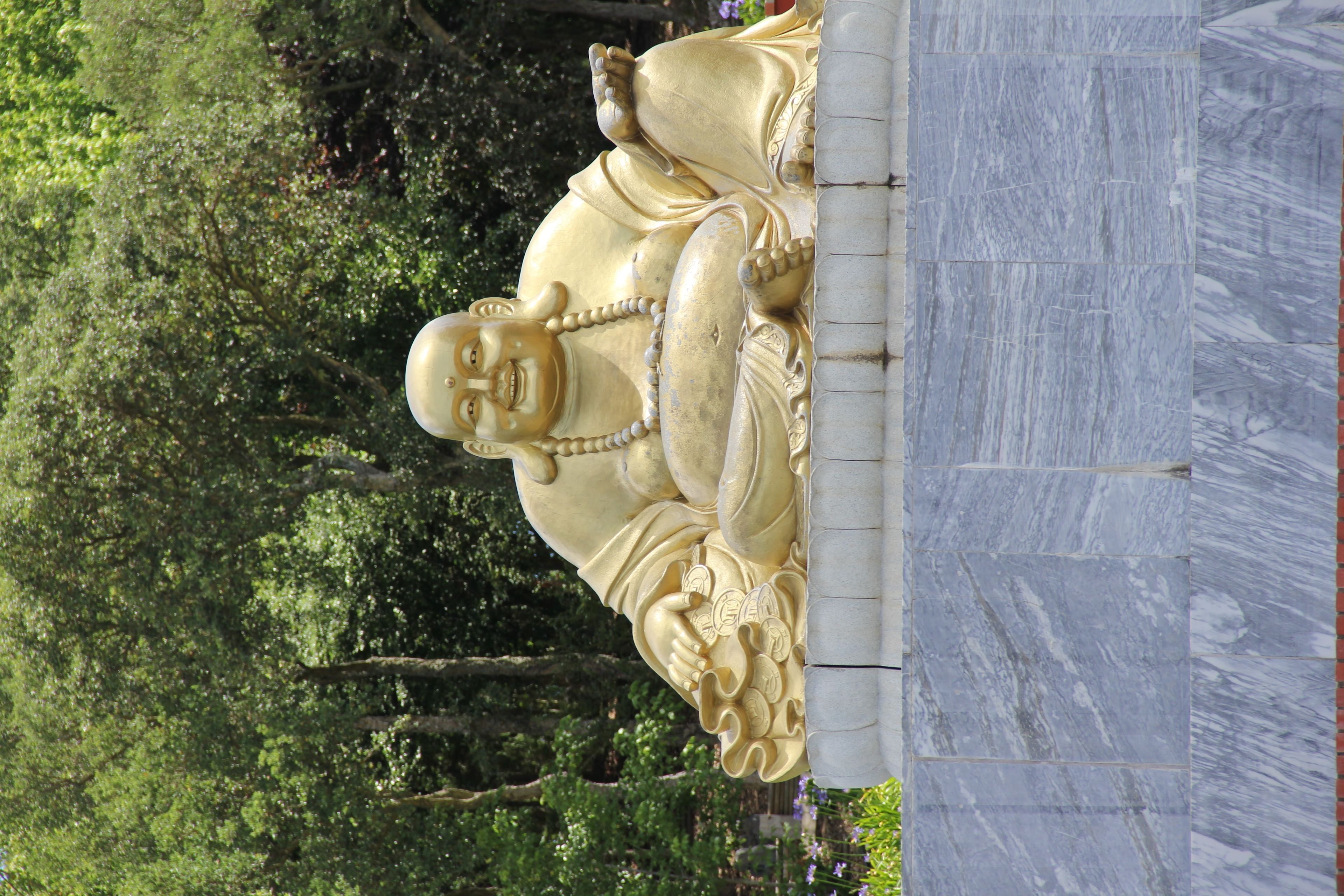 Una estatua dorada grande de una figura sentada con intrincados grabados, sobre un fondo de árboles verdes y frondosos.