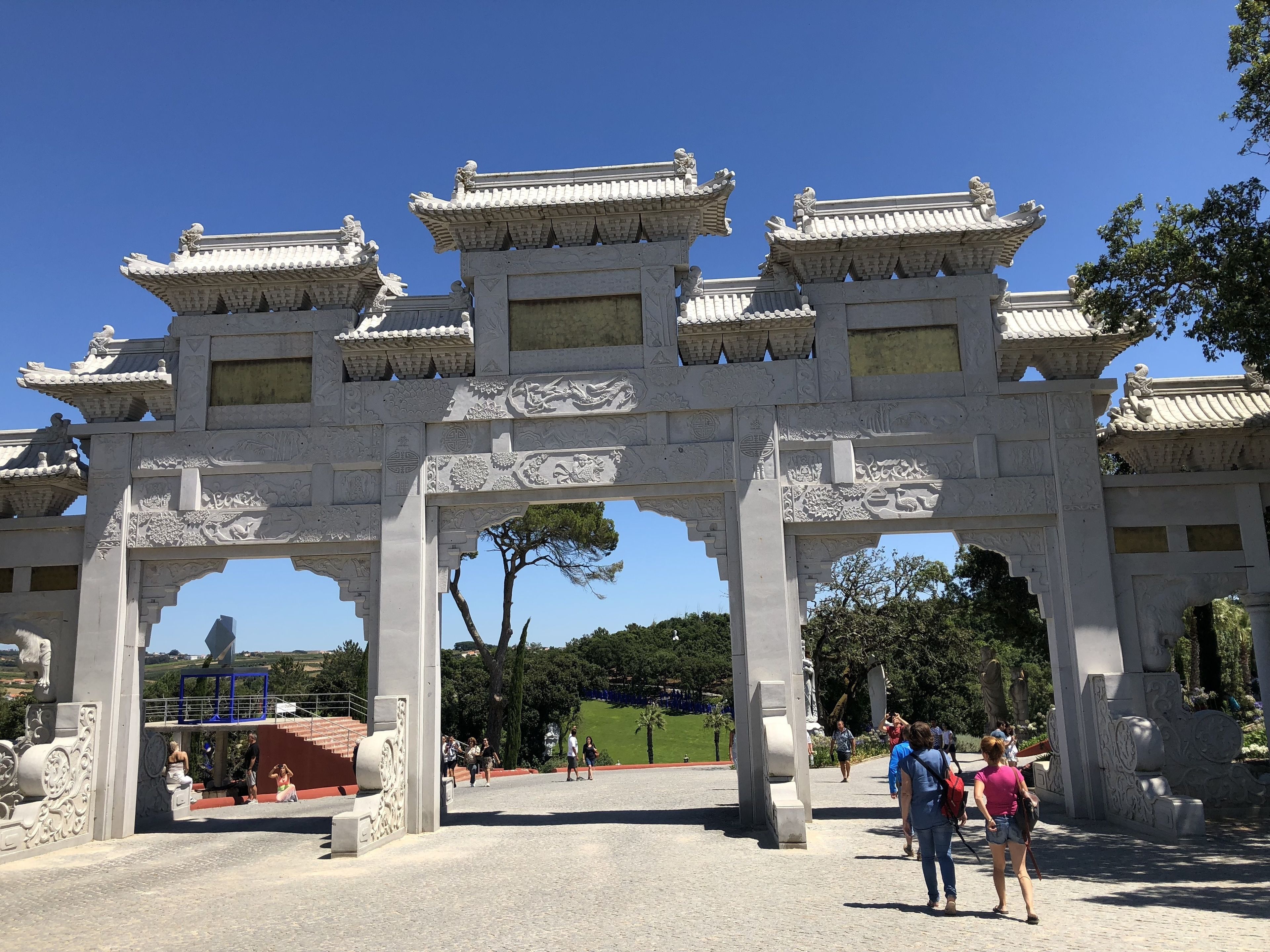 A traditional gate with intricate carvings and multiple tiers, leading to a park area with visitors.