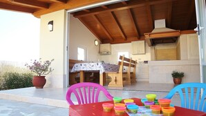 Une terrasse ouverte avec une table dressée pour le repas, un plafond en bois et un coin cuisine visible à travers la porte ouverte.