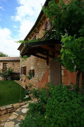 A stone building with a sloped roof, a covered porch, and a well-maintained garden with various plants and a lawn.