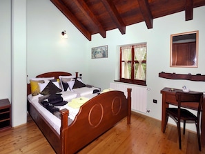 A bedroom with a wooden bed, a desk, a chair, a window with curtains, a radiator, and a picture on the wall.