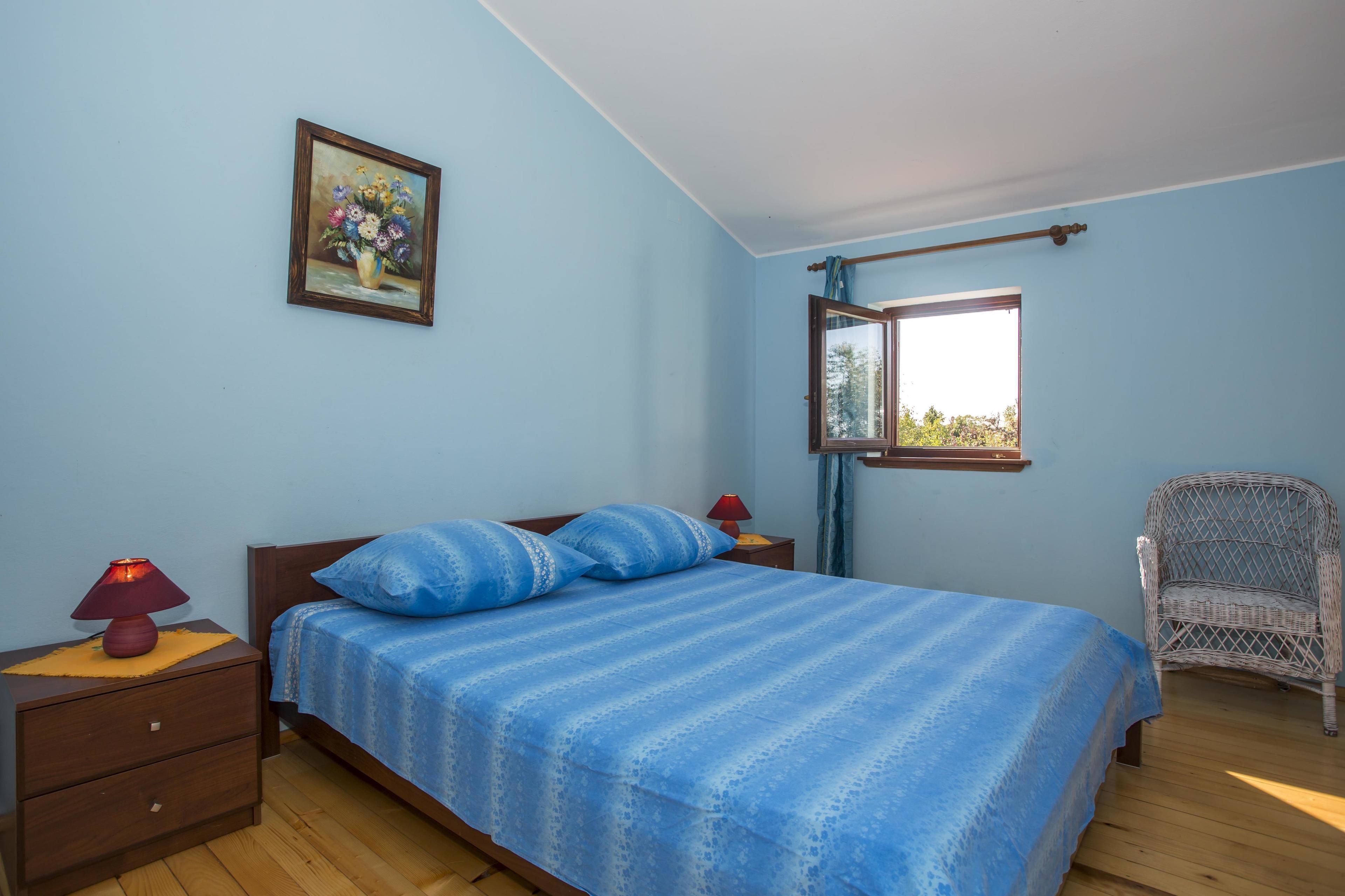 Een slaapkamer met een bed, nachtkastje, lamp, stoel, raam en een schilderij aan de muur.