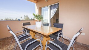 Une table en bois, six chaises, une plante en pot et une fenêtre avec des rideaux, le tout sur un balcon dont le sol est recouvert de carreaux.