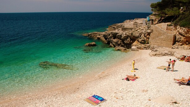 Een kiezelstrand met helder turquoise water, rotsachtige kliffen en enkele mensen die zonnebaden.