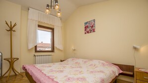 A bedroom with a bed, a window with curtains, a radiator, a coat rack, a bedside table, and a wall-mounted lamp.