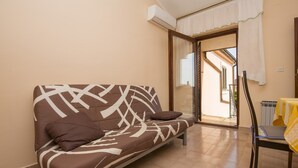 A room with a sofa, a chair, a table with a yellow cloth, and a sliding glass door leading to an outdoor area.