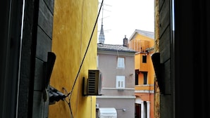 A window view of a narrow street with buildings on either side, including a yellow building with a black door and a brown building with a white awning. - Studio Erika (Rovinj)
