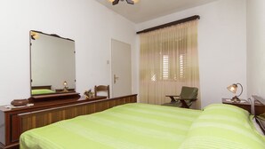 Ein ordentlich eingerichtetes Schlafzimmer mit einem großen Bett, einer hölzernen Kommode, einem Sessel, einem Schreibtisch mit Lampe und einem Fenster mit Vorhängen.
