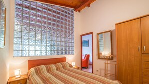 A bedroom with a large window, a bed with a striped bedspread, a nightstand with a lamp, a wooden wardrobe, and a mirror on the wall. - Apartment Nada / One Bedroom (Rovinj)