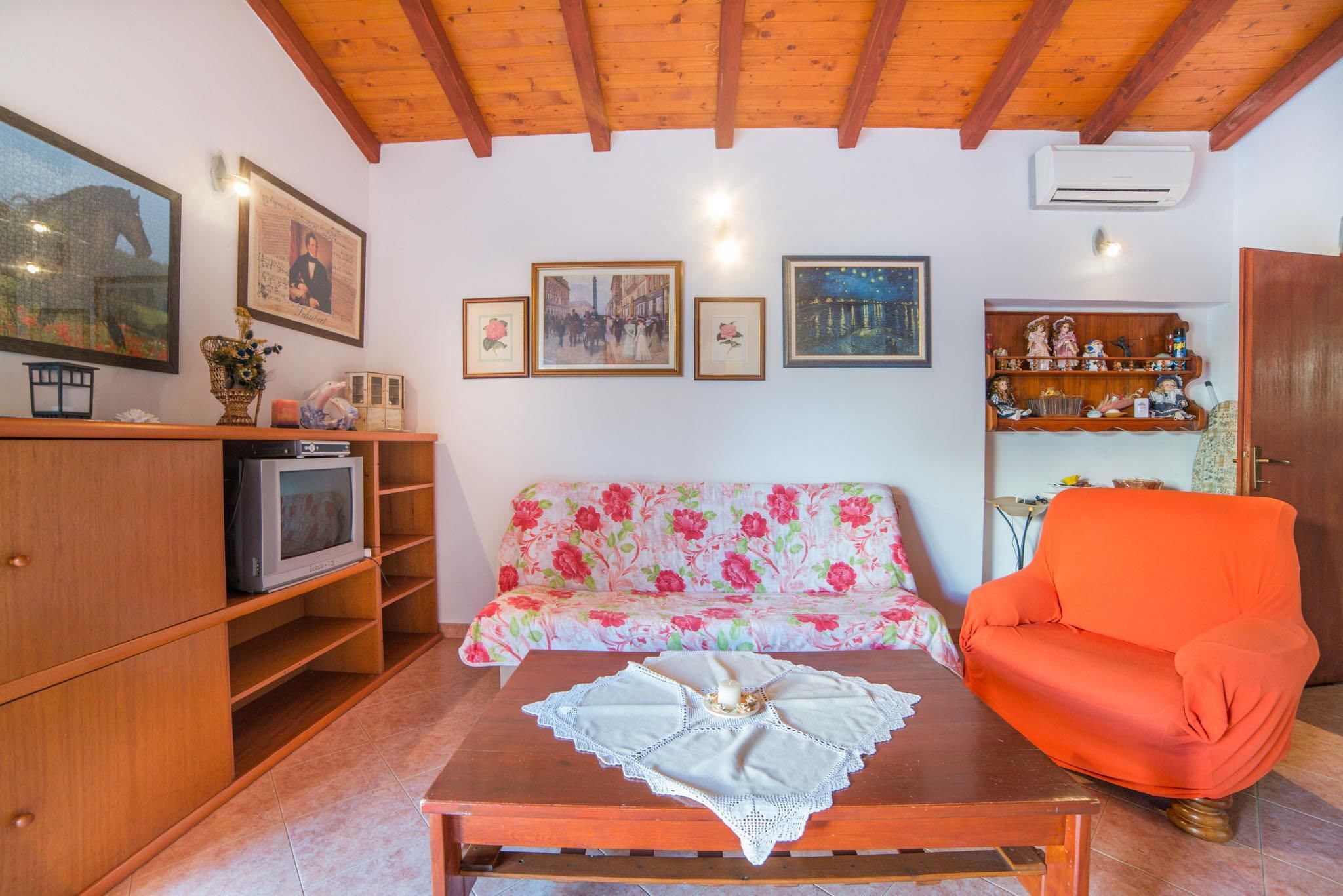 A cozy living room with a floral sofa, a wooden TV stand, a red armchair, and a wooden coffee table with a lace tablecloth.