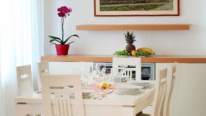 A dining area with a white table set for four, chairs, a red potted plant, and a framed painting above the table.