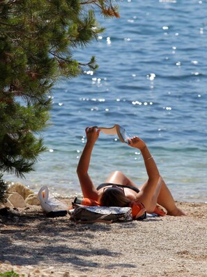 A person lying on a beach towel, reading a book, with a clear view of the sea and a pine tree in the background. - Apartments Biba Banjole / Two Bedrooms A1 (Medulin)