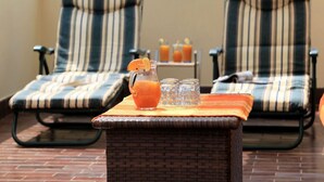 Two striped lounge chairs with a wicker table in between, a pitcher of orange juice, and a tray with two glasses.