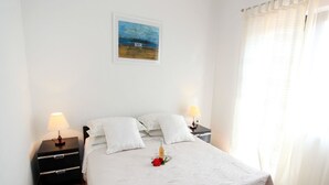 A neatly made bed with a white duvet, two bedside tables with lamps, a small bedside table with a bottle and a glass, a framed picture on the wall, and a window with sheer curtains.