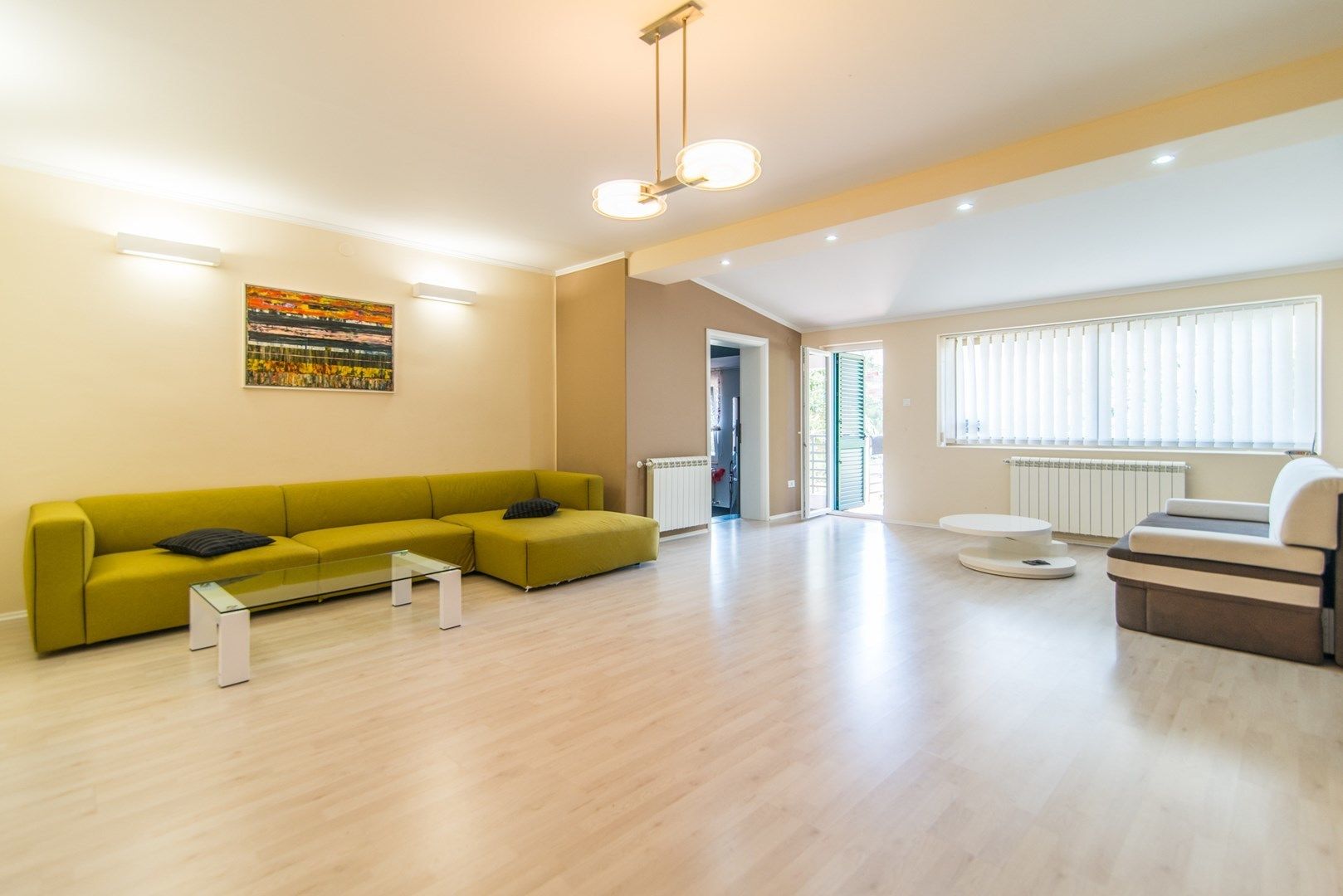 A modern living room with a green sofa, a glass coffee table, and a white armchair. There is a painting on the wall and a window with blinds.