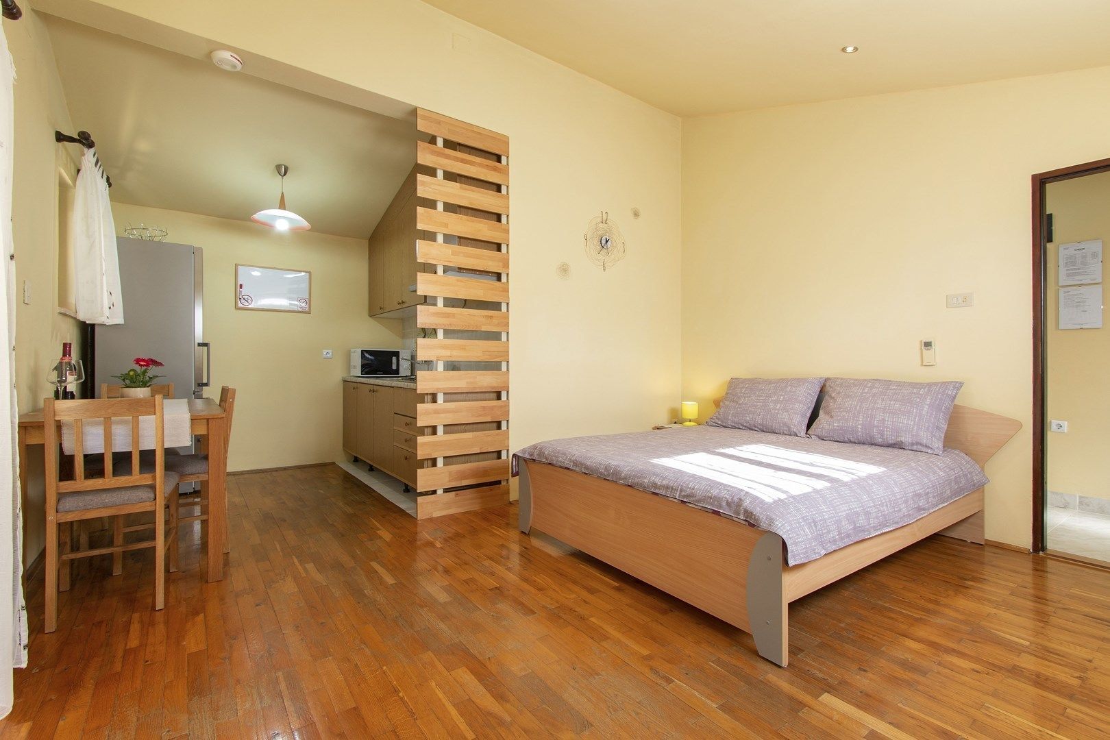 A bedroom with a wooden bed, a desk with a chair, a kitchenette, and a bathroom visible through an open door.