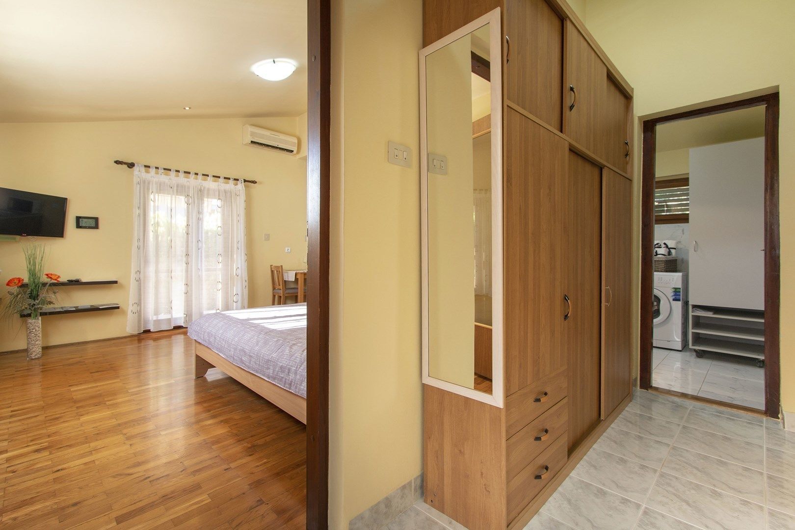 Een slaapkamer met een bed, een houten garderobe, een televisie en een wasmachine op de achtergrond.