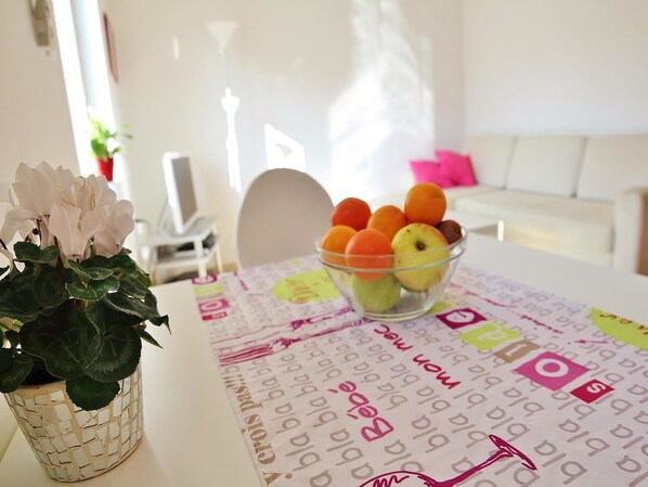 Una mesa con mantel blanco, letras y palabras del alfabeto, un bol de vidrio transparente con frutas y una planta en maceta con flores blancas.