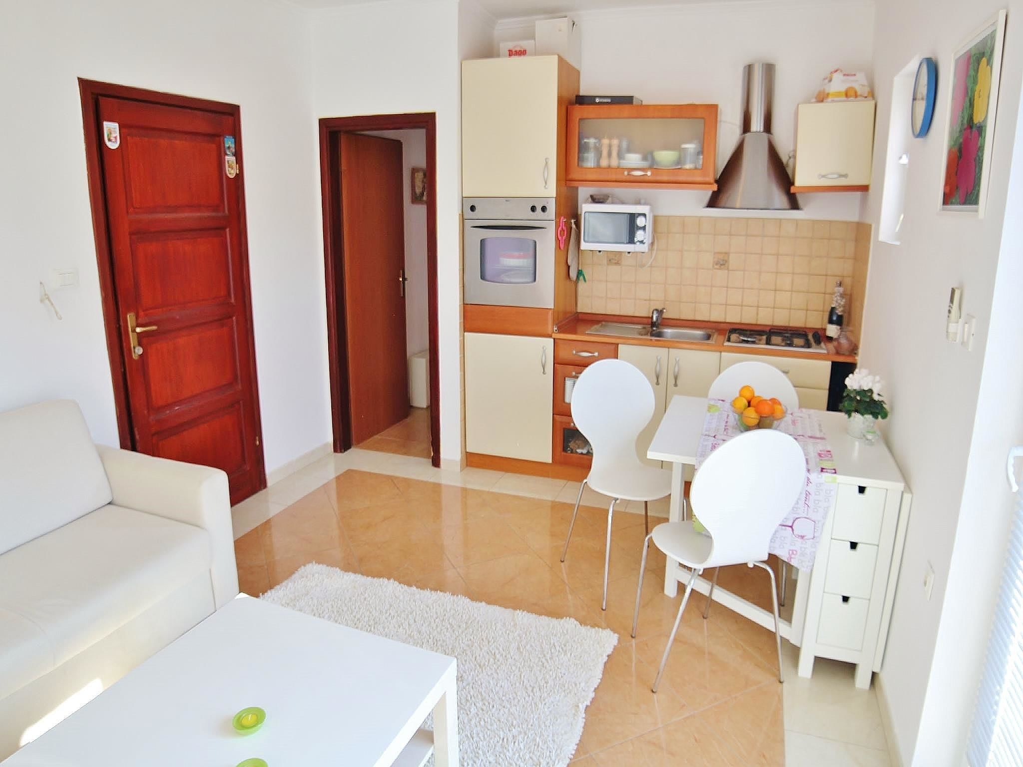 Cocina compacta con microondas, horno y armarios. Hay un comedor con mesa y sillas. En la sala de estar hay un sofá blanco y una mesita con alfombra.