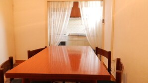 A dining area with a wooden table and chairs, a kitchen in the background, and sheer curtains on the windows.