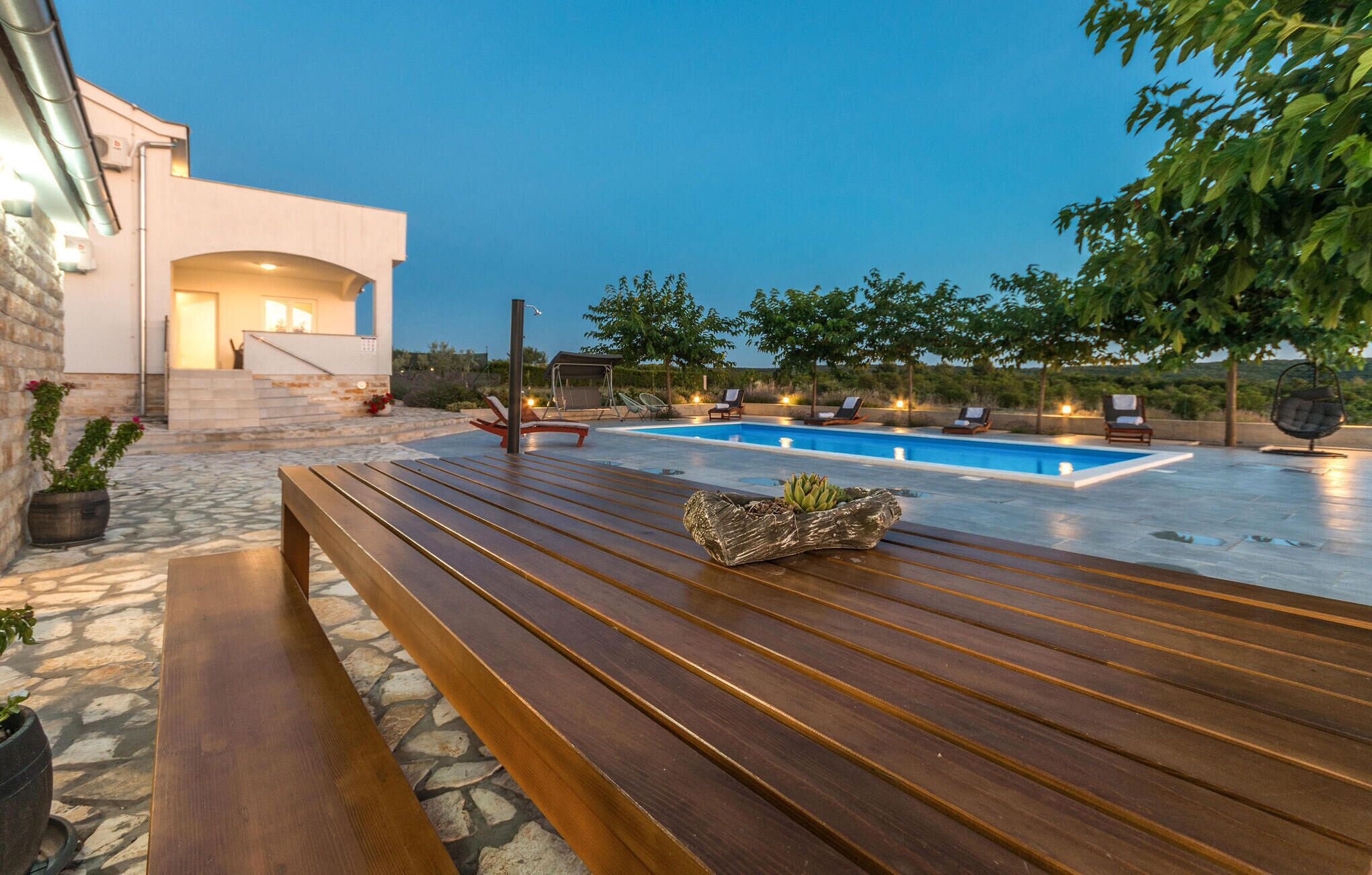 Un área moderna de piscina al aire libre con un banco de madera, sillas de descanso y un sendero de piedra que lleva a un edificio con una escalera.