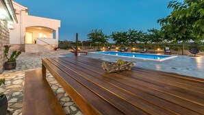 Uma área moderna com piscina ao ar livre, um banco de madeira, cadeiras de descanso e um caminho de pedra que leva a um edifício com escadaria.