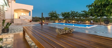 Un área moderna de piscina al aire libre con un banco de madera, sillas de descanso y un sendero de piedra que lleva a un edificio con una escalera.