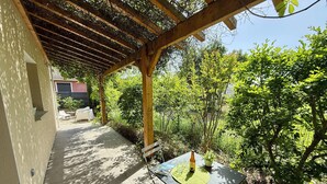 A covered patio area with a table and chairs, surrounded by lush greenery and a view of a house in the background.