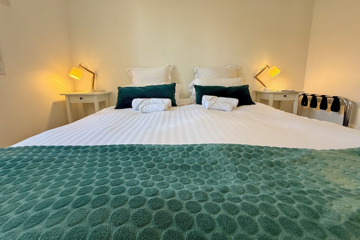 A neatly made bed with a green textured bedspread and white pillows, two bedside lamps, and a headboard with a towel rack.