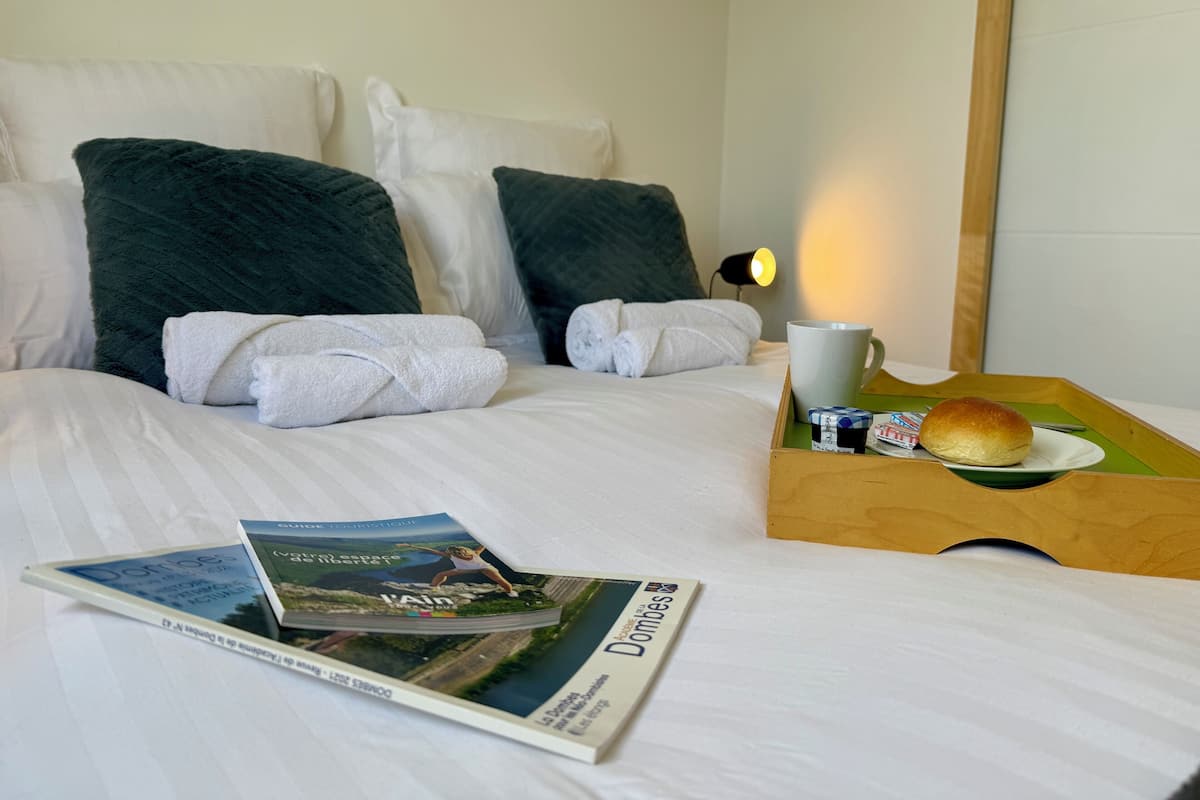 A neatly made bed with white linens, two dark green pillows, and a pair of white towels. A wooden tray holds a white mug, a small plate with a bun, and a magazine.