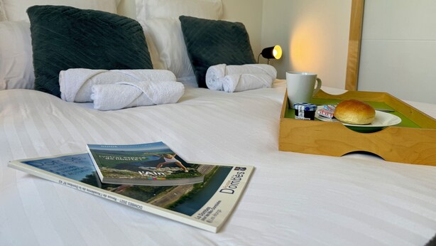 A neatly made bed with white linens, two dark green pillows, and a pair of white towels. A wooden tray holds a white mug, a small plate with a bun, and a magazine.
