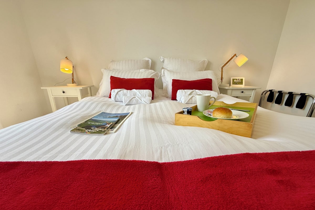 A neatly made bed with white and red bedding, a magazine, a tray with food and a mug, and bedside lamps.