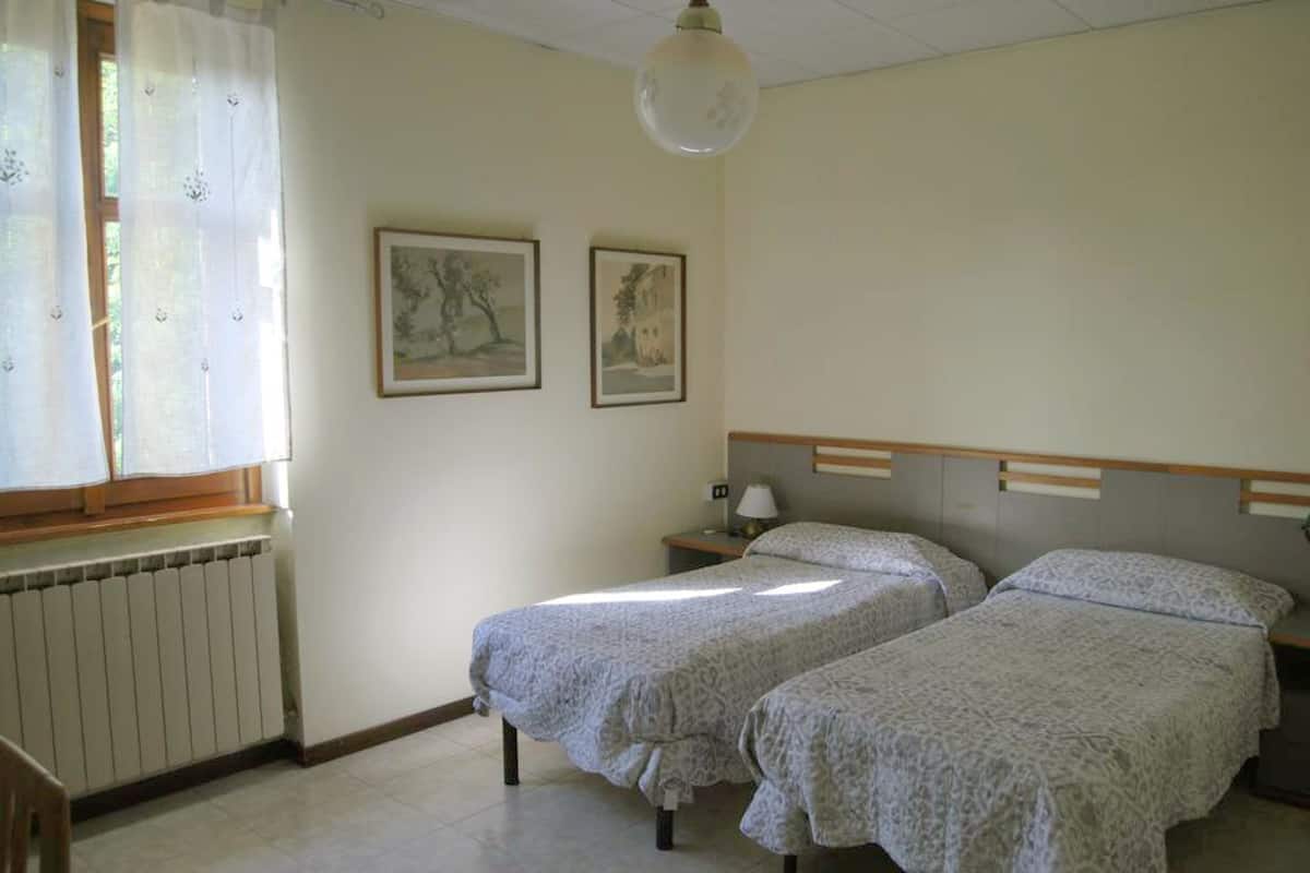 A room with two beds, a radiator, a window with curtains, and two framed pictures on the wall.