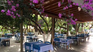 Un espacio de comedor al aire libre con mesas y sillas bajo una pérgola de madera, rodeado de árboles y follaje.