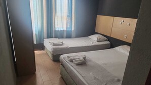 Habitación de hotel con dos camas, cabecero de madera, un mueble pequeño y una ventana con cortinas translúcidas.