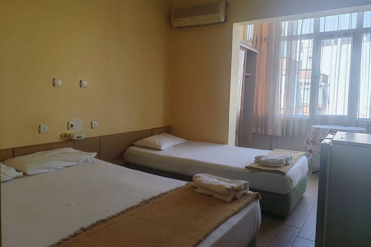 A hotel room with two single beds, a small desk, a window with curtains, and an air conditioning unit on the wall.