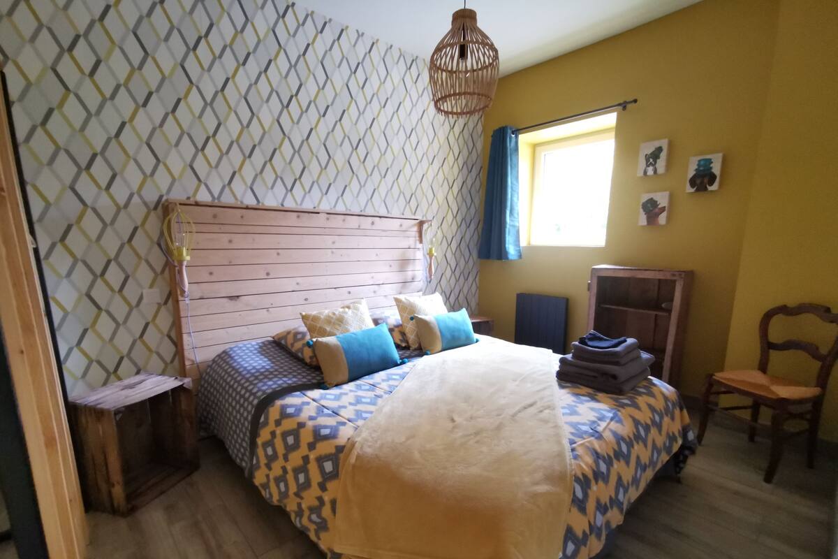 A bedroom with a wooden headboard, patterned bedding, a small bedside table, a chair, and a window with curtains.