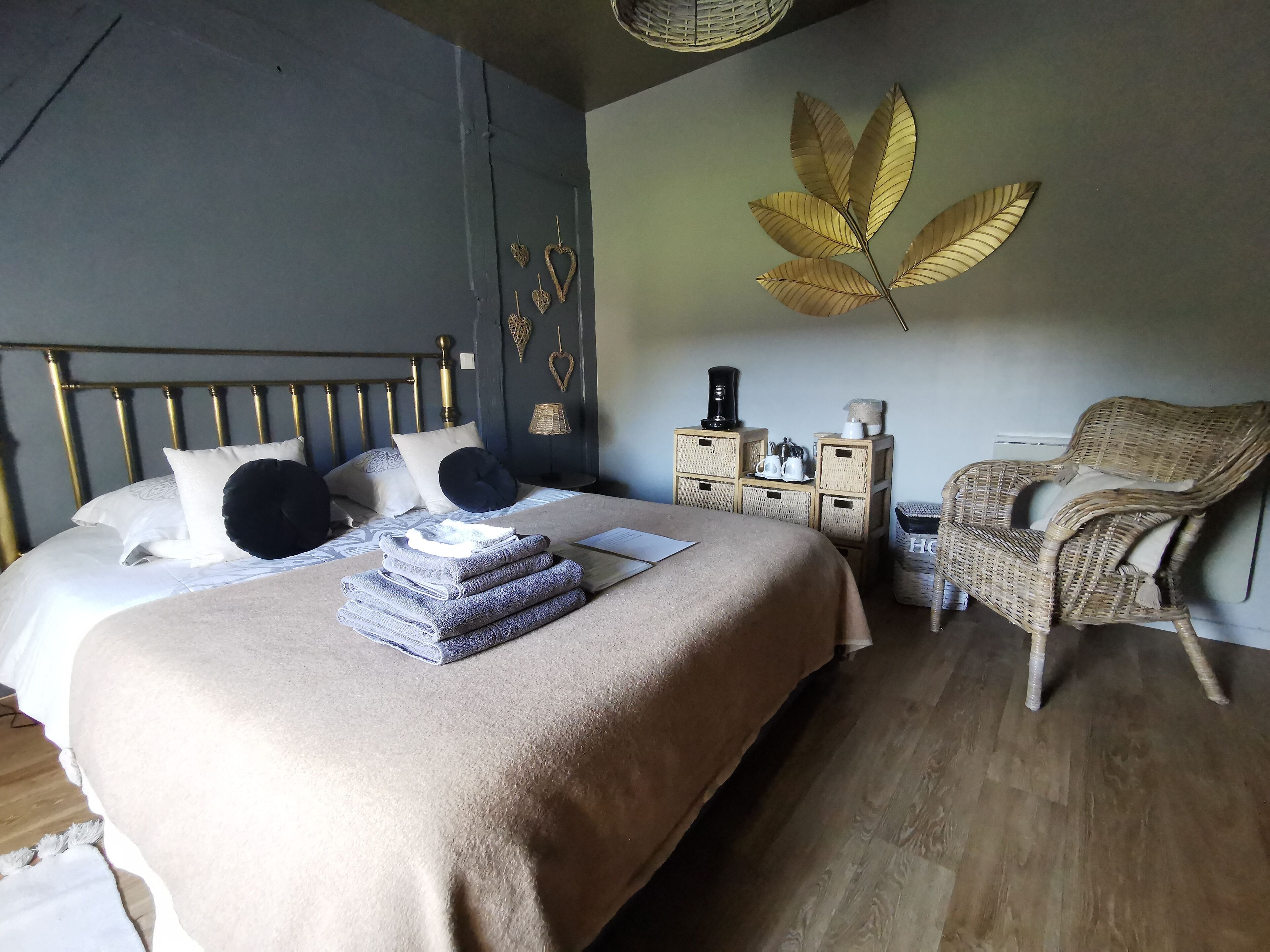 Una cama hecha con orden, cubierta con un edredón color beige, una pila de toallas dobladas y una almohada negra. Se observa una silla de mimbre y una mesita con una lámpara y una cafetera.