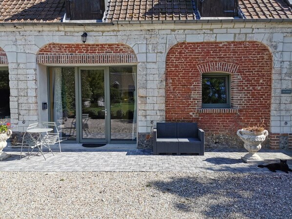 A building with a brick facade, a glass door entrance, outdoor seating, and a small window. - La Cour d'Hortense (Sailly-Flibeaucourt)