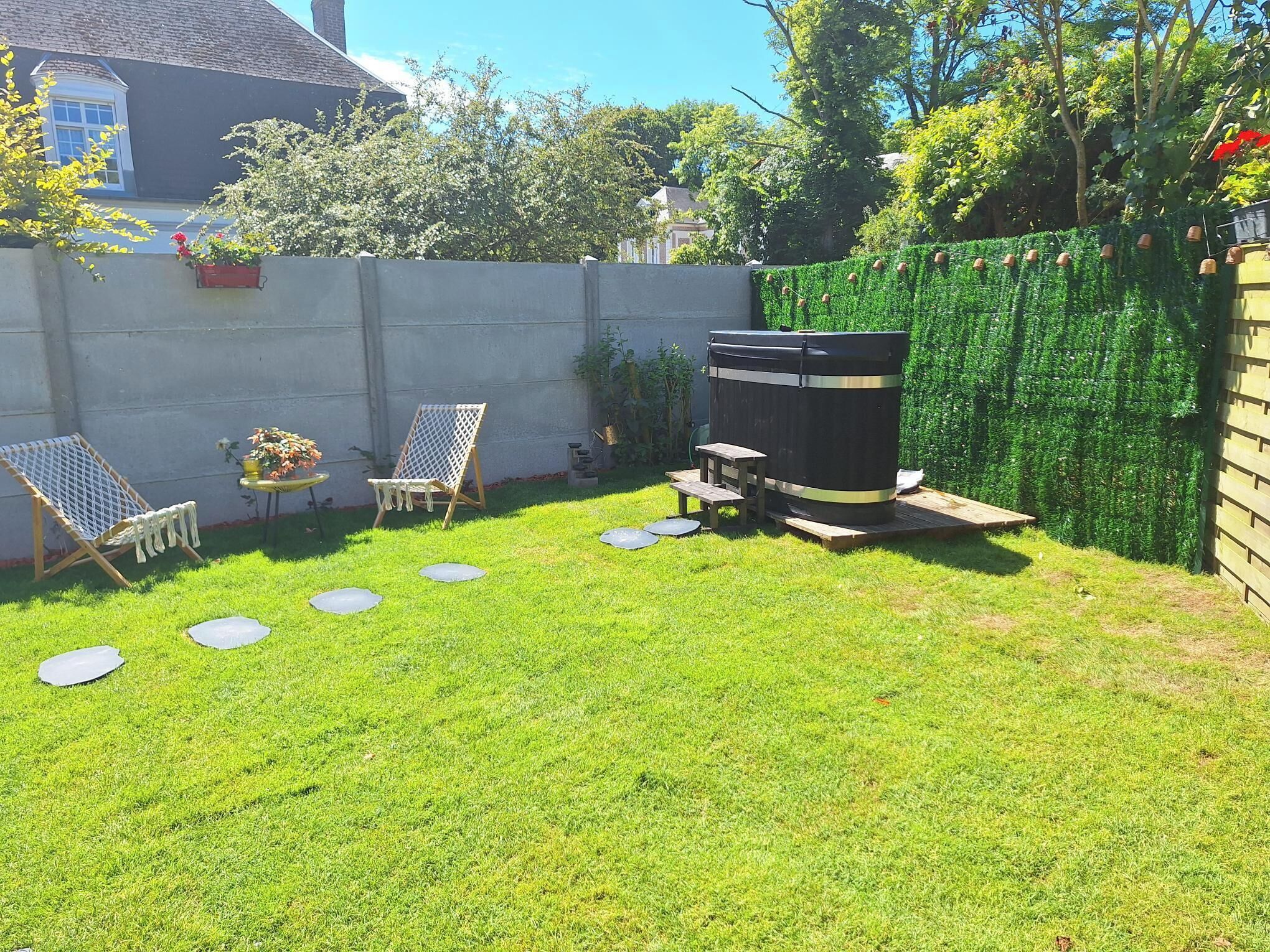 A backyard with a barbecue grill, two chairs, a small table, and a garden area with a hedge.
