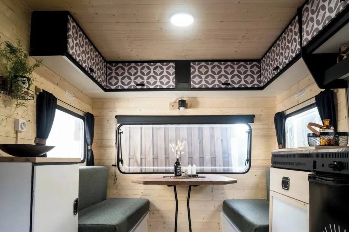 A compact RV interior with a wooden table, two seating areas, a window with curtains, and a kitchenette.