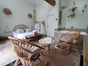 A well-lit room with a bed, a wooden table, wicker chairs, a small round table, and a bathroom with a bathtub.