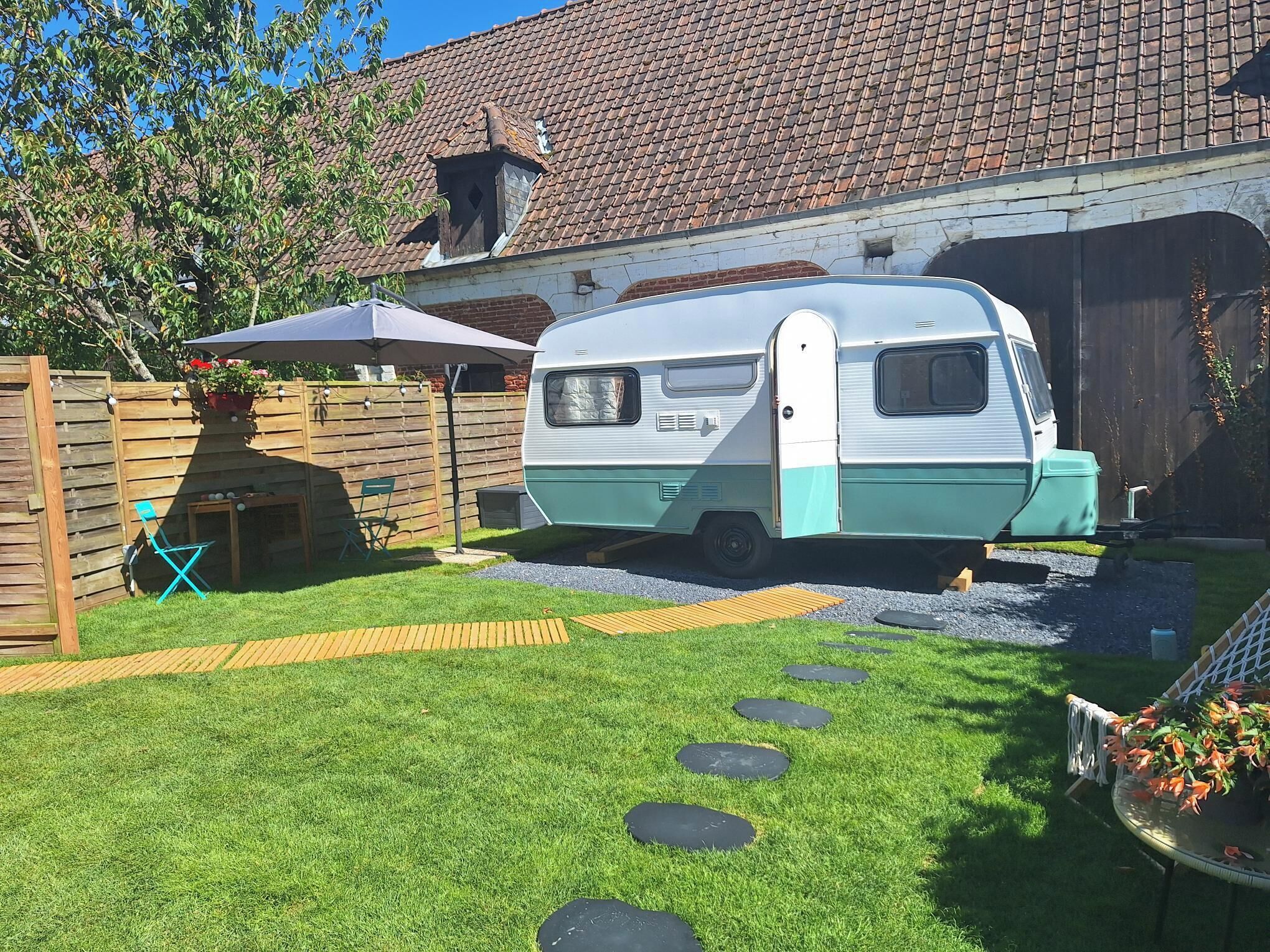 A caravan parked in a backyard with a grassy lawn, stepping stones, a wooden fence, and a tiled roof.