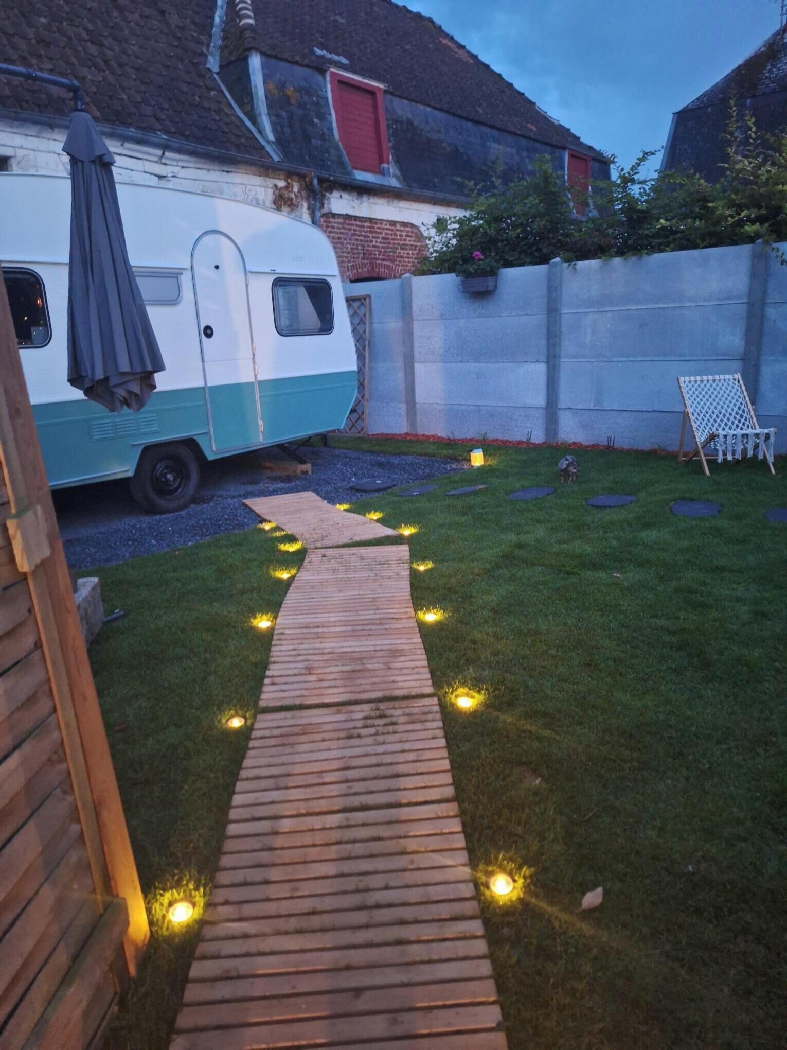 Un patio trasero con un sendero de madera, una caravana blanca y azul, y una silla blanca.