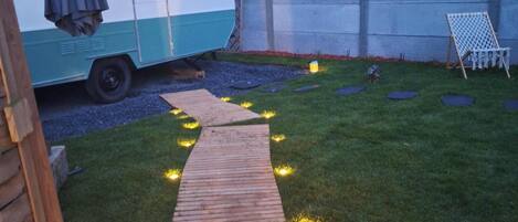 A backyard with a wooden pathway, a white and blue caravan, and a white chair.